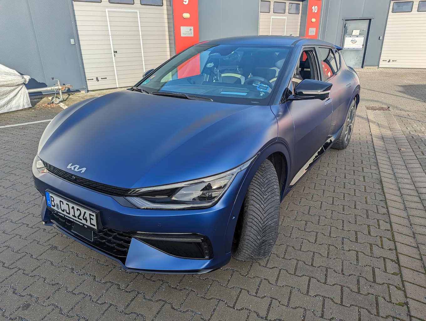 Ein blauer Kia EV6 steht schräg auf einer gepflasterten, grauen Fläche. Das linke Vorderrad steht nach außen. Das Fahrefenster ist offen. Die Fronthaube ist präsent nach vorn gerichtet.